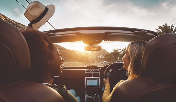 Two ladies in a car singing along to the music as they drive into the sunset